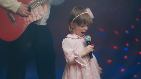 Little girl on stage in vintage dress, she sings into microphone and dances, her father plays acoustic guitar. Color music is shining and smoke is billowing. Father and Child Day, performance on stageの写真素材
