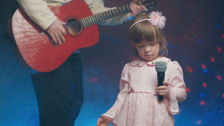 Little girl on stage in vintage dress, she sings into microphone and dances, her father plays acoustic guitar. Color music is shining and smoke is billowing. Father and Child Day, performance on stageの写真素材