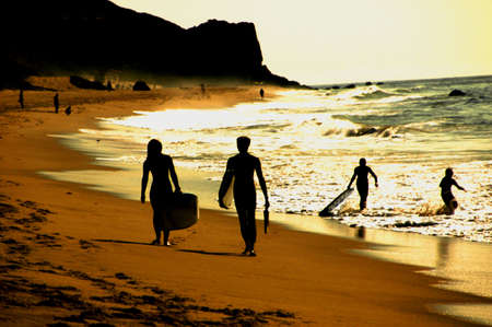 Silhouette of two surfers walking on beach, people playing in background.の写真素材