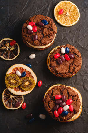 Homemade oat chocolate cookies sandwich with dried citrus fruits and juicy jelly beans on textured dark black background, top viewの写真素材