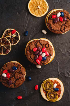 Homemade oat chocolate cookies sandwich with dried citrus fruits and juicy jelly beans on textured dark black background, top viewの写真素材