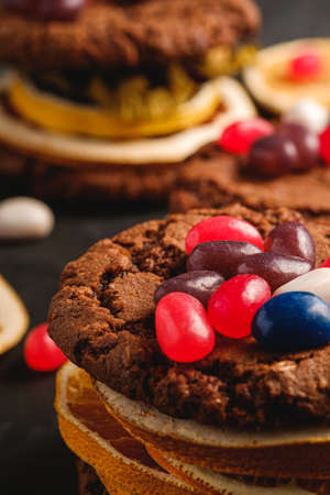Homemade oat chocolate cookies sandwich with dried citrus fruits and juicy jelly beans on textured dark black background, angle view macroの写真素材