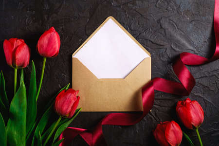 Red bunch of tulip flowers with envelope card and ribbon on textured black background, top view copy...