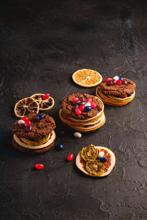 Homemade oat chocolate cookies sandwich with dried citrus fruits and juicy jelly beans on textured dark black background, angle viewの写真素材