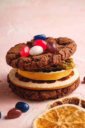 Homemade oat chocolate cookies sandwich with dried citrus fruits and juicy jelly beans on textured pink background, angle viewの写真素材