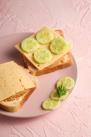 Two fresh square sandwiches with turkey ham meat, cheese, greenery dill and cucumber slices on pink plate on textured background, angle viewの写真素材