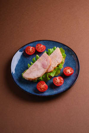 Sandwich with turkey ham meat, green salad and fresh cherry tomatoes slices on blue shiny plate, brown minimal background, angle viewの写真素材