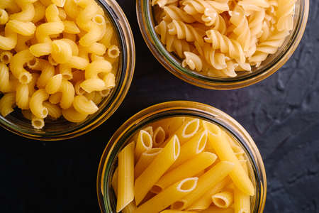 Three glass jars with variety of uncooked golden wheat pasta on dark black textured background, top view macroの写真素材