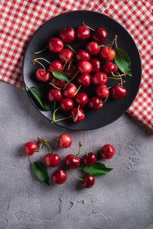 Fresh ripe cherry fruits with green leaves on black plate with red dish towel, summer vitamin berries on grey stone background, top viewの写真素材