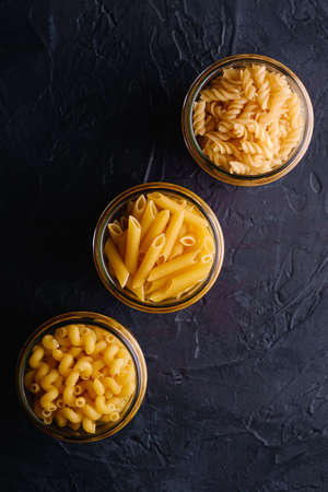 Three glass jars in row with variety of uncooked golden wheat pasta on dark black textured background, top viewの写真素材