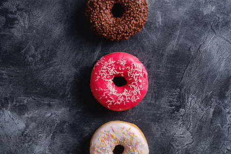 Chocolate, pink and vanilla donuts with sprinkles in row, sweet glazed dessert food on dark concrete textured background, top viewの写真素材