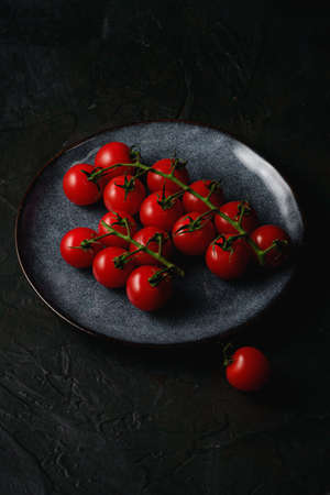 Cherry tomato branch, fresh ripe organic vegetables in grey plate on dark black textured background, angle viewの写真素材