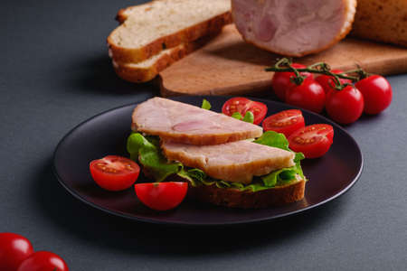 Sandwich with turkey ham meat, green salad and fresh cherry tomatoes slices on black plate near to ingredients on cutting board, grey minimal background, angle view macroの写真素材