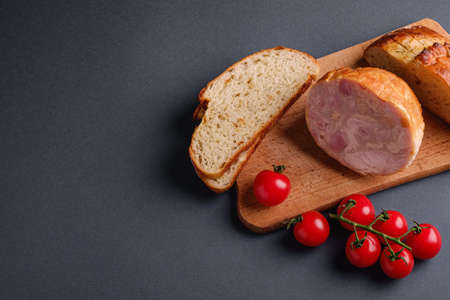 Turkey ham meat, fresh cherry tomatoes branch and loaf sliced bread on wooden cutting board, grey minimal background, angle view, copy spaceの写真素材
