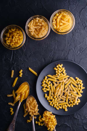 Three cutlery spoons, glass jars and plate with variety of uncooked golden wheat pasta on dark black textured background, top viewの写真素材