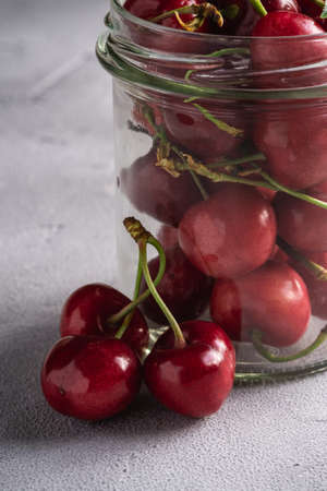 Fresh ripe cherry branch near to cherries with green leaves in glass jar, summer vitamin berries on grey stone background, angle view macroの写真素材