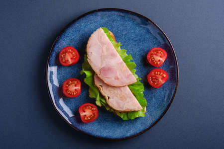 Sandwich with turkey ham meat, green salad and fresh cherry tomatoes slices on blue shiny plate, blue minimal background, top viewの写真素材