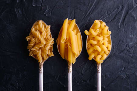 Three cutlery spoons with variety of uncooked golden wheat pasta on dark black textured background, top viewの写真素材