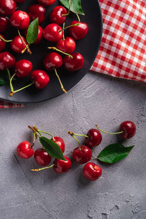 Fresh ripe cherry fruits with green leaves on black plate with red dish towel, summer vitamin berries on grey stone background, top viewの写真素材