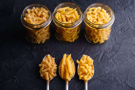 Three cutlery spoons and glass jars with variety of uncooked golden wheat pasta on dark black...