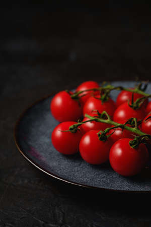 Cherry tomato branch, fresh ripe organic vegetables in grey plate on dark black textured background, angle viewの写真素材