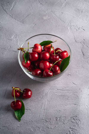 Fresh ripe cherry fruits with green leaves in glass bowl, summer vitamin berries on grey stone background, angle viewの写真素材