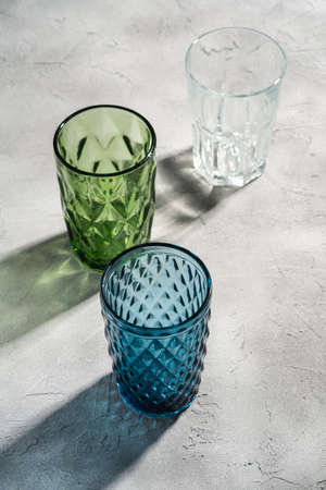 Three glass geometric cup in blue, green and transparent colors with colorful shadow light rays on stone concrete background, angle viewの写真素材