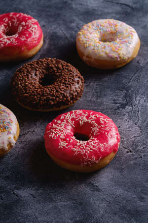 Chocolate, pink and vanilla donuts with sprinkles, sweet glazed dessert food on dark concrete textured background, angle viewの写真素材