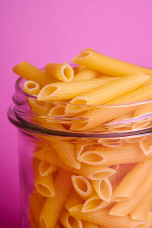 One glass jar with penne uncooked golden wheat tube pasta on minimal pink background, angle view macroの写真素材
