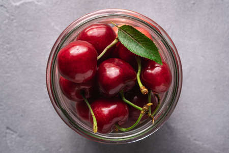 Fresh ripe cherry fruits with green leaves in glass jar, summer vitamin berries on grey stone background, top view macroの写真素材