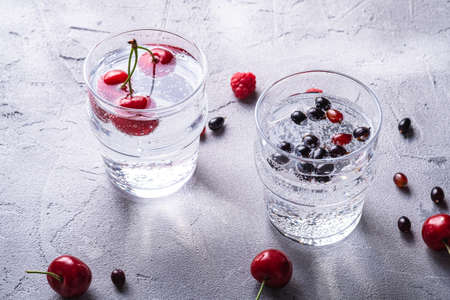 Fresh cold sparkling water drink with cherry, raspberry and currant berries in two transparent glass on stone concrete background, summer diet beverage, angle viewの写真素材