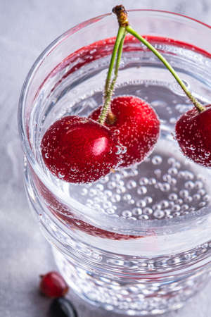 Fresh cold sparkling water drink with cherry, raspberry and currant berries in transparent glass on stone concrete background, summer diet beverage, angle view macroの写真素材