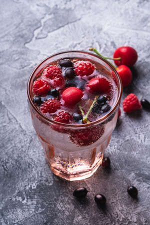 Fresh cold sparkling water drink with cherry, raspberry and currant berries in red faceted glass on stone concrete background, summer diet beverage, angle viewの写真素材