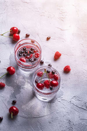 Fresh cold sparkling water drink with cherry, raspberry and currant berries in two transparent glass on stone concrete background, summer diet beverage, angle viewの写真素材