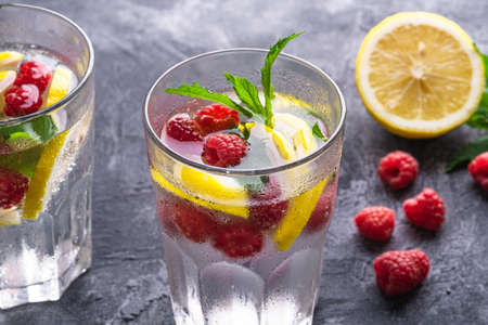 Fresh cold ice water drink with lemon, raspberry fruits and mint leaf in two faceted glass on stone concrete background, summer diet beverage, angle view selective focusの写真素材