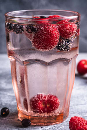 Fresh cold sparkling water drink with cherry, raspberry and currant berries in red faceted glass on stone concrete background, summer diet beverage, angle view macroの写真素材