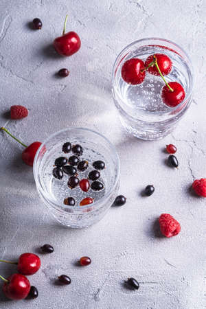 Fresh cold sparkling water drink with cherry, raspberry and currant berries in two transparent glass on stone concrete background, summer diet beverage, angle viewの写真素材