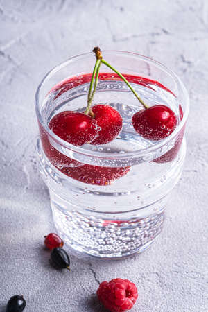 Fresh cold sparkling water drink with cherry, raspberry and currant berries in transparent glass on stone concrete background, summer diet beverage, angle view macroの写真素材