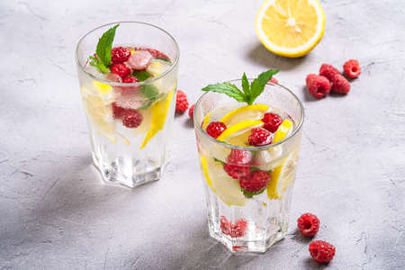 Fresh cold ice water drink with lemon, raspberry fruits and mint leaf in two faceted glass on stone concrete background, summer diet beverage, angle viewの写真素材