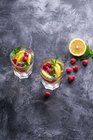 Fresh cold ice water drink with lemon, raspberry fruits and mint leaf in two faceted glass on stone concrete background, summer diet beverage, angle view selective focusの写真素材
