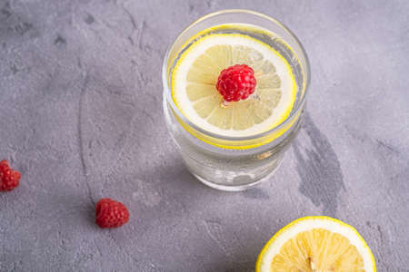Fresh cold sparkling water drink with lemon, raspberry fruits in glass on stone concrete background, summer diet beverage, angle view macroの写真素材
