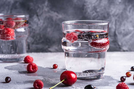 Fresh cold sparkling water drink with cherry, raspberry and currant berries in two transparent glass on stone concrete background, summer diet beverage, angle view macroの写真素材