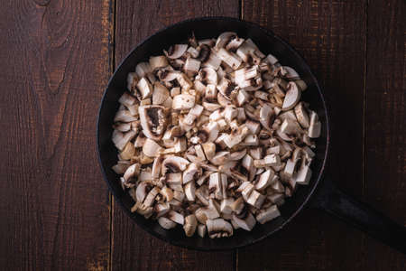 Tasty chopped champignon mushrooms in frying pan, uncooked food sliced, dark brown wooden background, top viewの写真素材