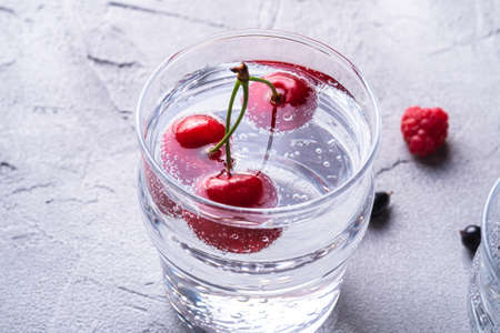 Fresh cold sparkling water drink with cherry, raspberry and currant berries in transparent glass on stone concrete background, summer diet beverage, angle view macroの写真素材