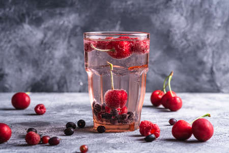 Fresh cold sparkling water drink with cherry, raspberry and currant berries in red faceted glass on stone concrete background, summer diet beverage, angle viewの写真素材