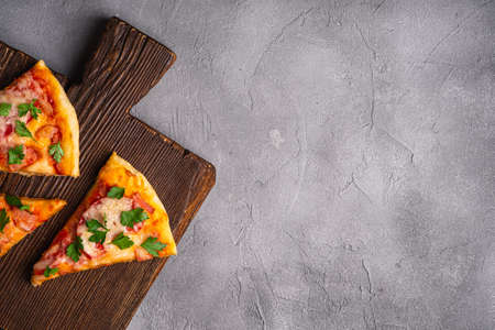 Hot pizza slices with mozzarella cheese, ham, tomato and parsley on brown wooden cutting board, stone concrete background, top view copy space for textの写真素材