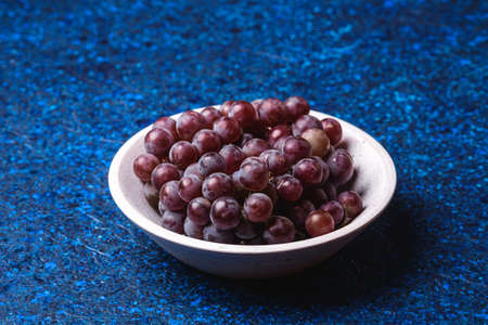 Fresh ripe grape berries in white wooden bowl on blue abstract background, angle viewの写真素材
