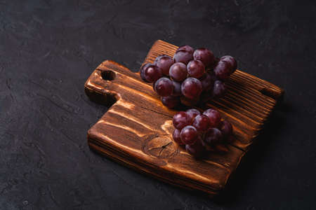 Fresh ripe grape berries on brown wooden cutting board on dark stone background, angle viewの写真素材