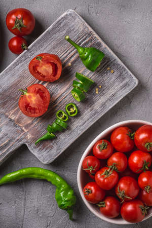 Fresh ripe tomatoes in bowl near to old wooden cutting board with hot chili peppers, stone concrete background, top viewの写真素材
