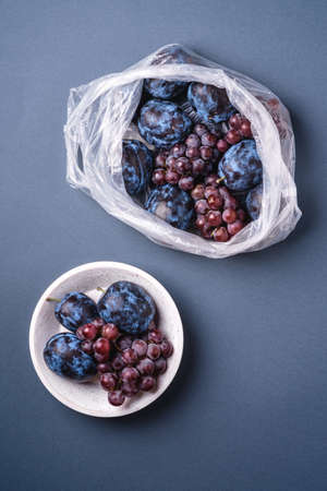 Fresh ripe plum fruits and grape berries in plastic bag package and in wooden bowl on minimal blue grey background, top viewの写真素材